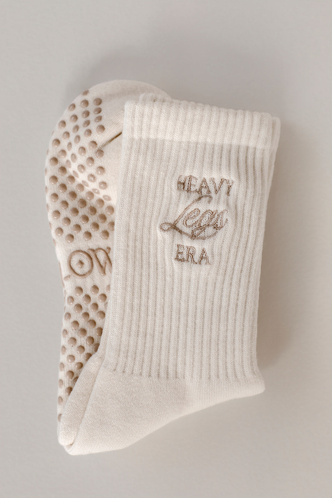 Light colored sock with textured grip sole and embroidered text on a neutral background. Socks say Heavy Legs Era.