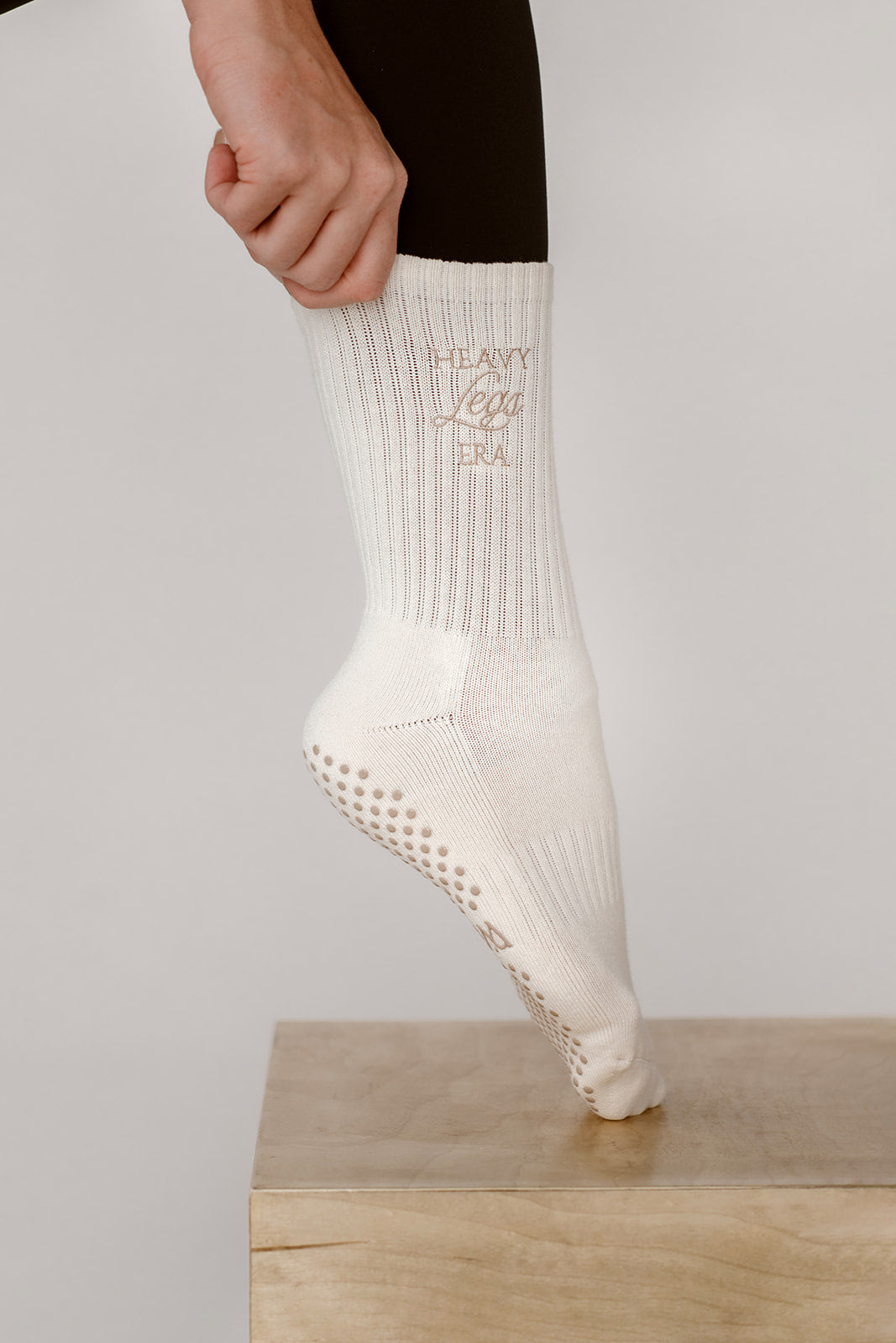 White sock with taupe text worn by a person on a light gray background. Socks say Heavy Legs Era.